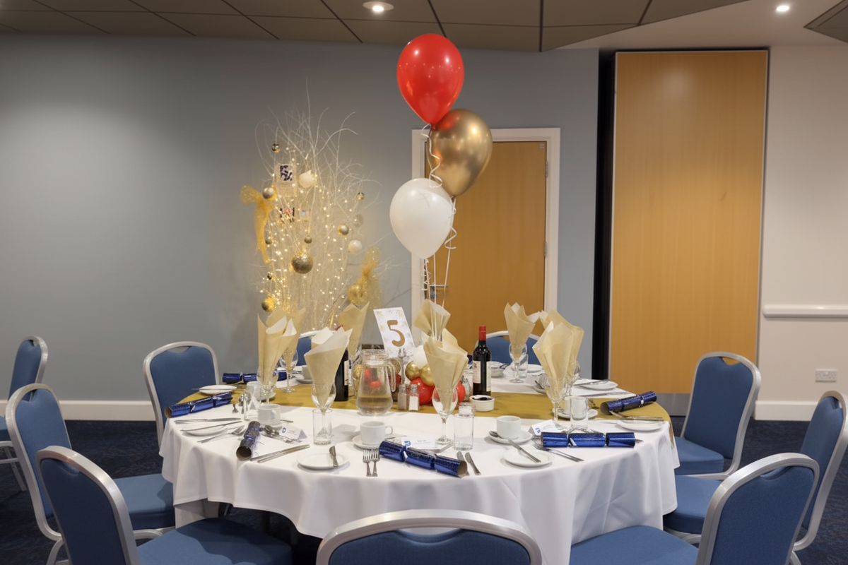 A decorated round banquet table with a white tablecloth, plates, cutlery, and gold and white centerpieces, featuring red, gold, and white balloons in a conference room.
